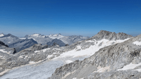 A high-altitude panorama with rocky peaks and small glaciers as far as the eye can see, all under a bright blue sky