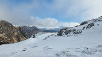 a Scottish mountain landscape with snow in the foreground and sunlit snow-free rocky slopes on a hillside at the left of frame. The sky is patchy with clouds